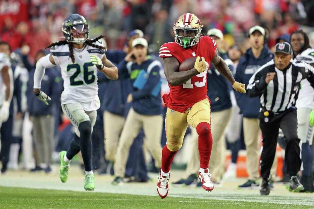 jogador do 49ers carrega bola com jogador do seahawks correndo ao fundo no wild card da NFL 2022-23