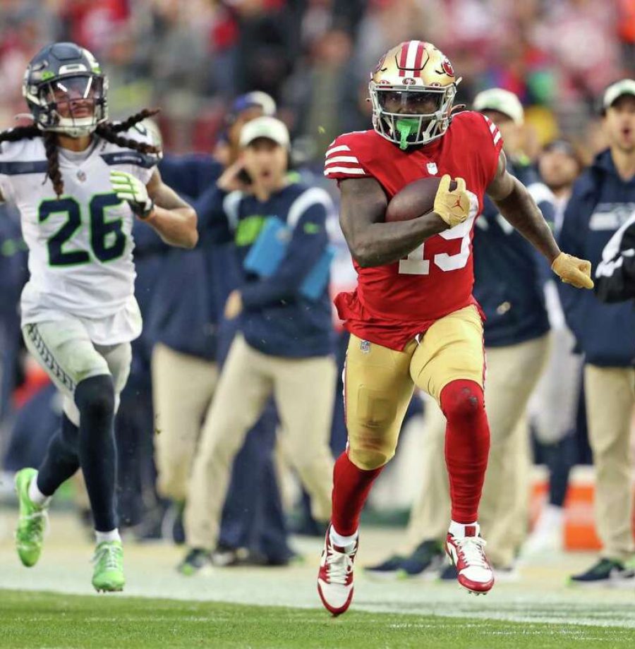 jogador do 49ers carrega bola com jogador do seahawks correndo ao fundo no wild card da NFL 2022-23